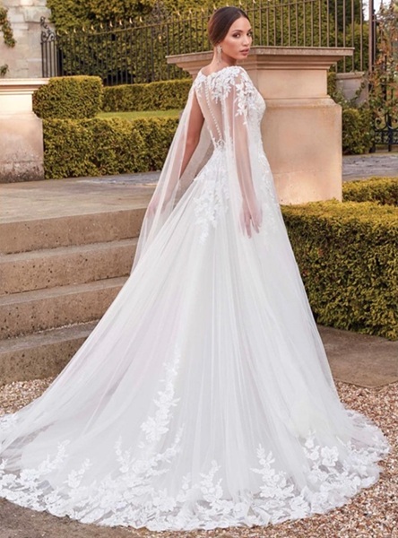Sposa di spalle in abito bianco di tulle con pizzo floreale, mantella trasparente e strascico in un giardino con siepi