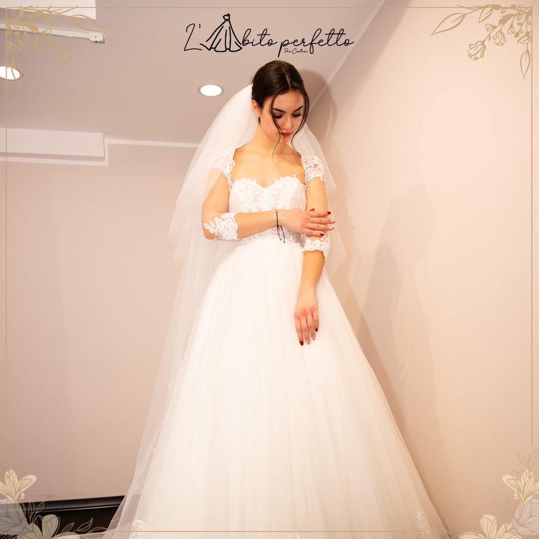 Sposa con velo e abito bianco in tulle, corpetto e maniche in pizzo, nell'atelier con logo L'Abito Perfetto