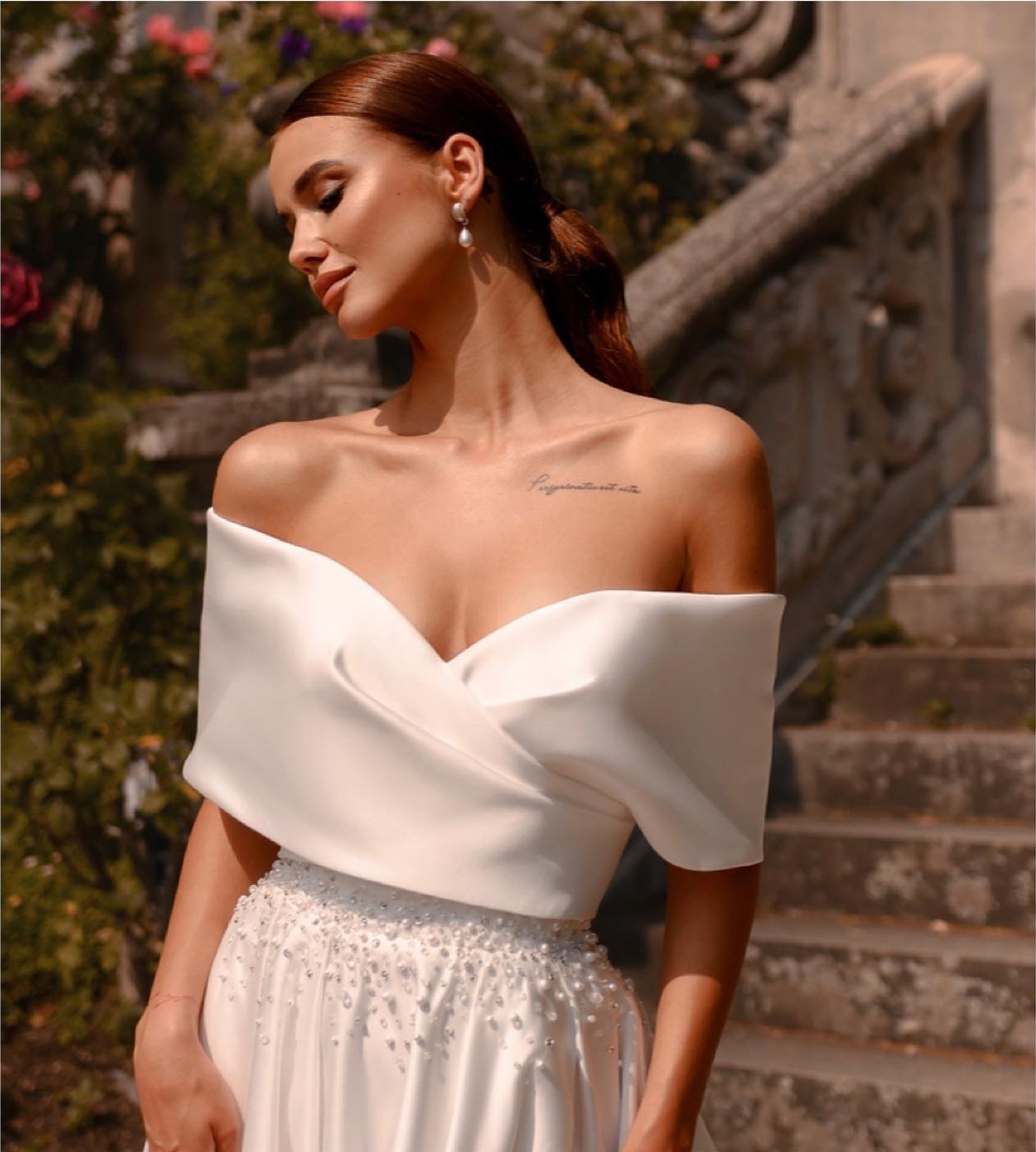 Sposa in abito bianco con scollo incrociato off-shoulder e gonna di perline, di L'Abito Perfetto, davanti a una scala in pietra